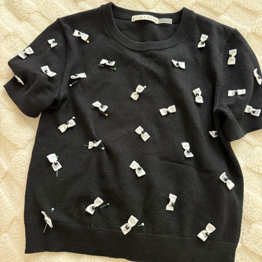 Black knit sweater with scattered white fabric bows, laid flat on cream background — After Stilettos alteration repair