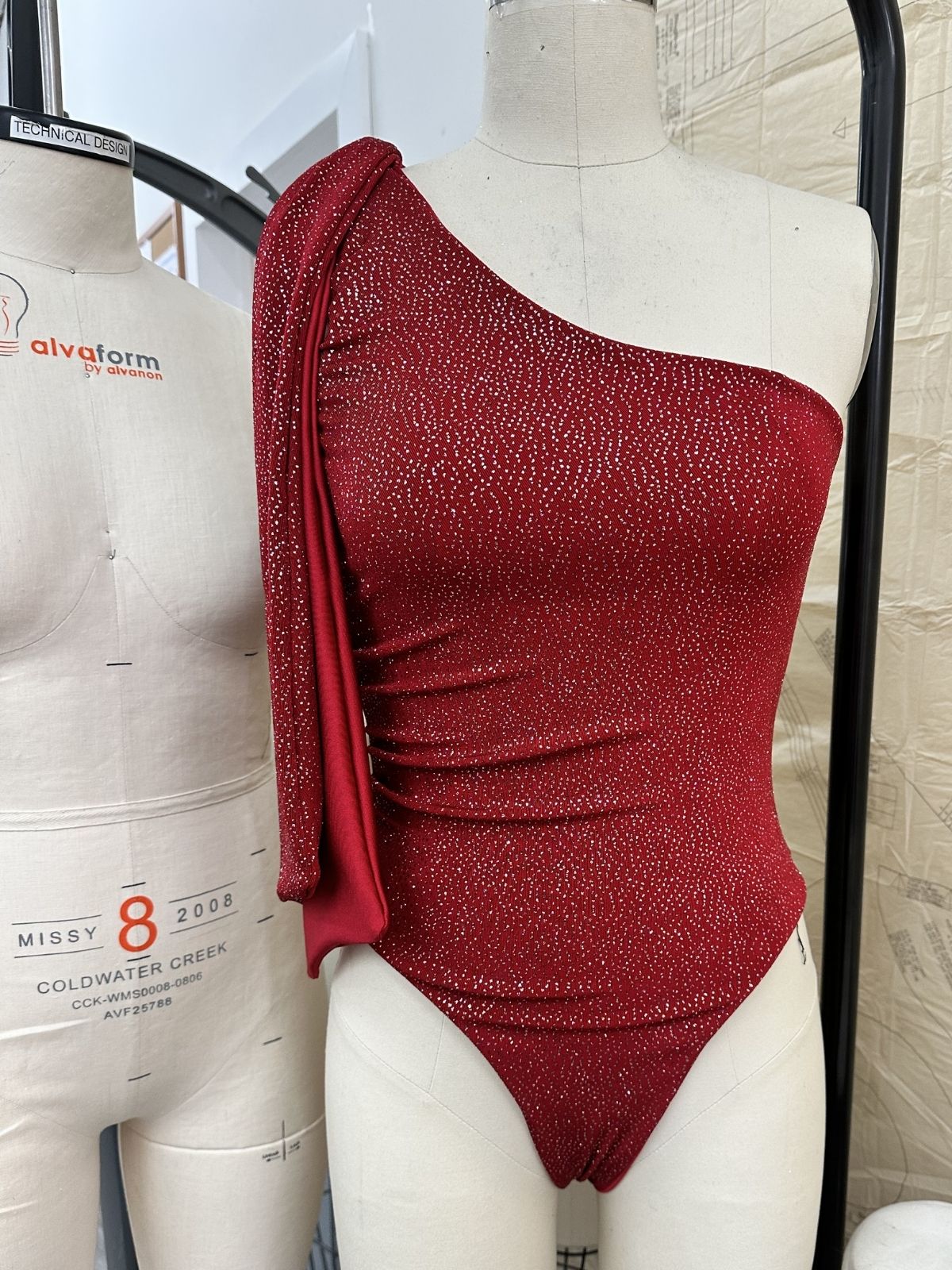 After Stilettos Crimson Sculpt one-shoulder sequin one-piece swimsuit on dress form showing draped sleeve construction and ruched body
