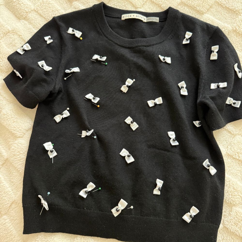 Black knit sweater with scattered white fabric bows, laid flat on cream background — After Stilettos alteration repair

