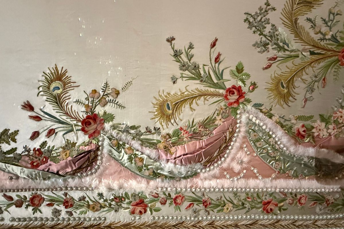 Close-up of an elaborately embroidered 18th century bodice with silk roses, peacock feathers, and pearl trim on display at the Marie Antoinette exhibition, V&A London
